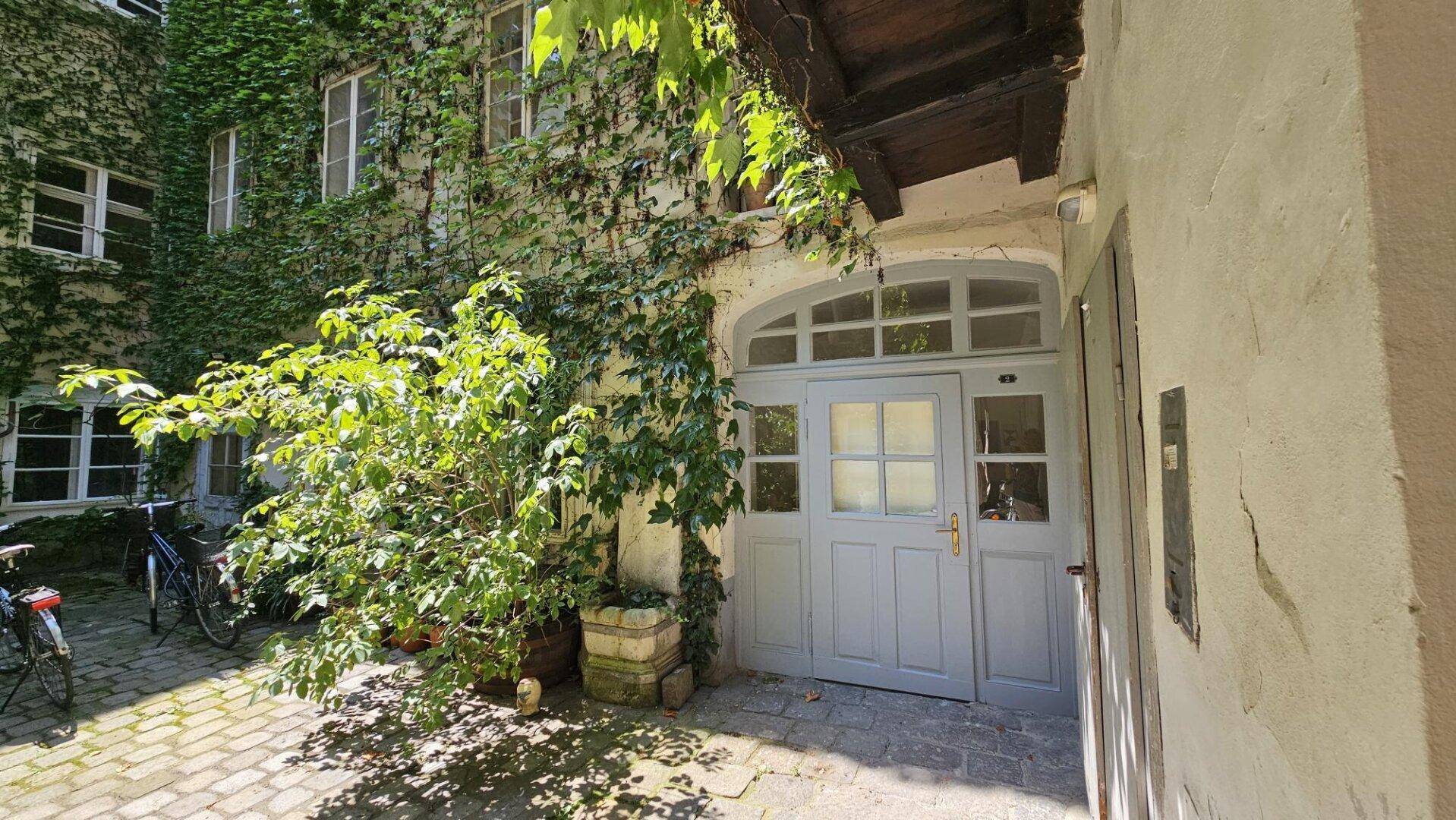 Historischer Eingang mit grauer Doppeltür im begrünten Innenhof, umgeben von Efeu und Pflanzen.