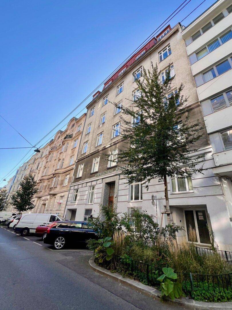 Straßenansicht eines Altbaus mit Bäumen, parkenden Autos und angrenzenden Gebäuden.