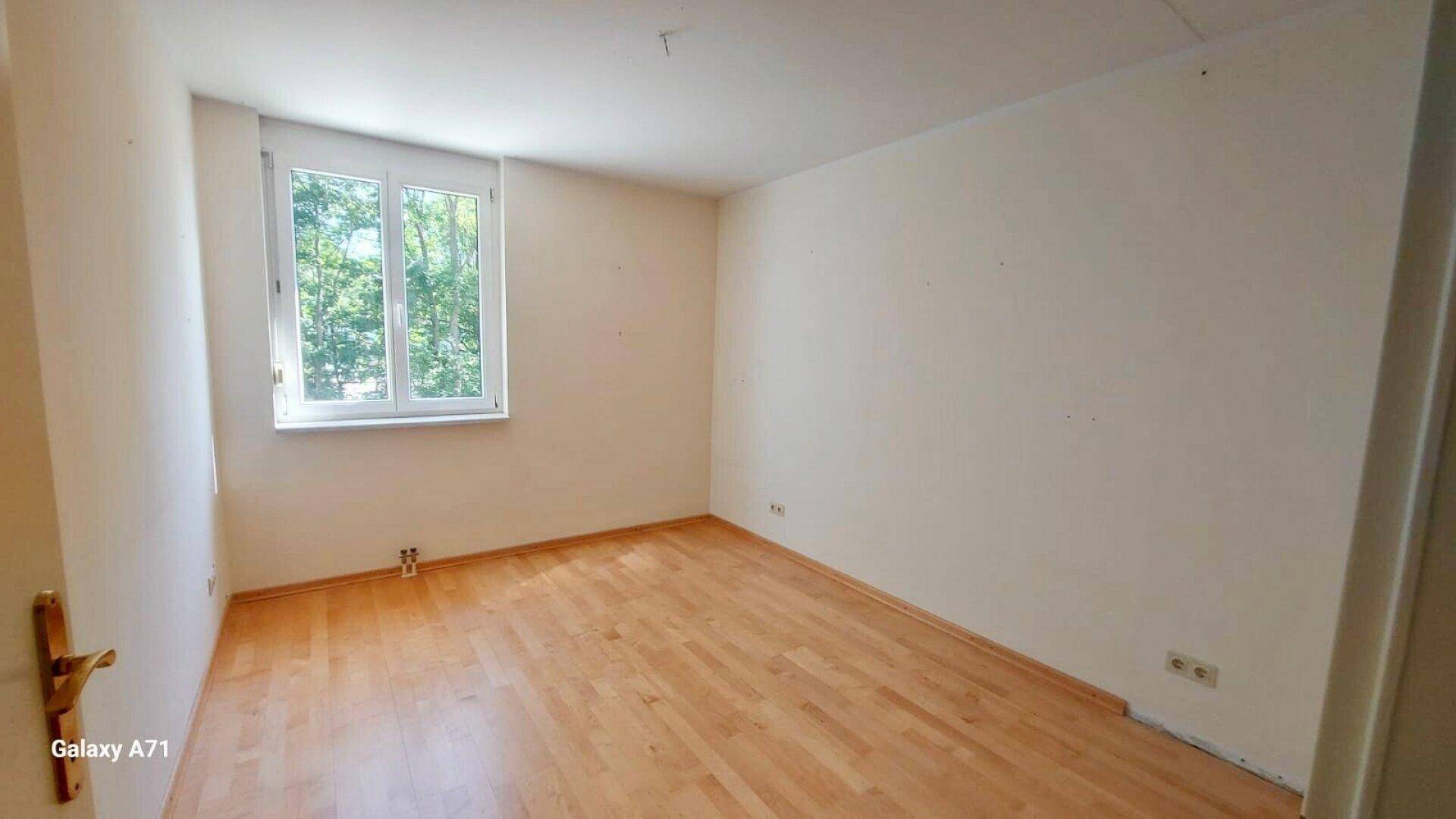 Schlafzimmer mit hellem Holzparkettboden und einem Fenster mit Blick ins Grüne.