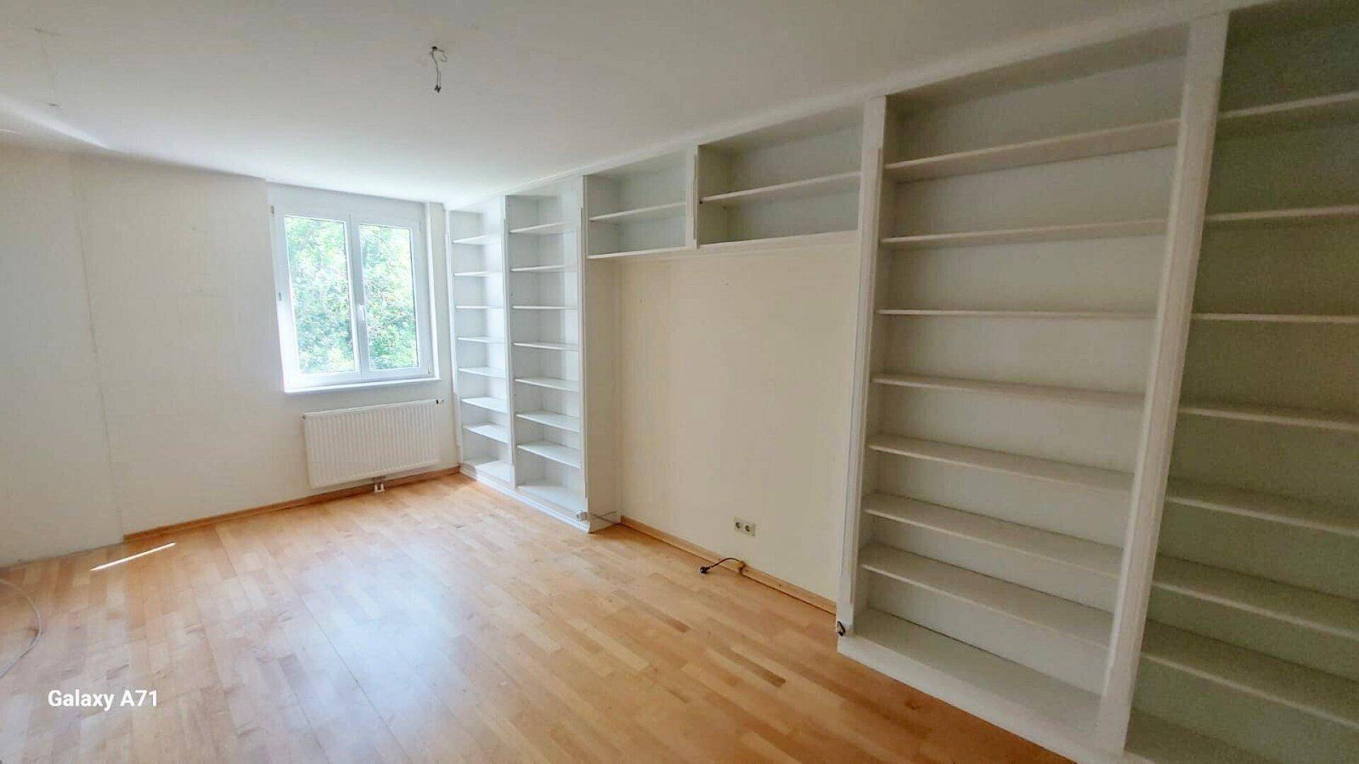 Helles Zimmer mit Holzparkett und umfangreichen Einbauregalen entlang der Wände.