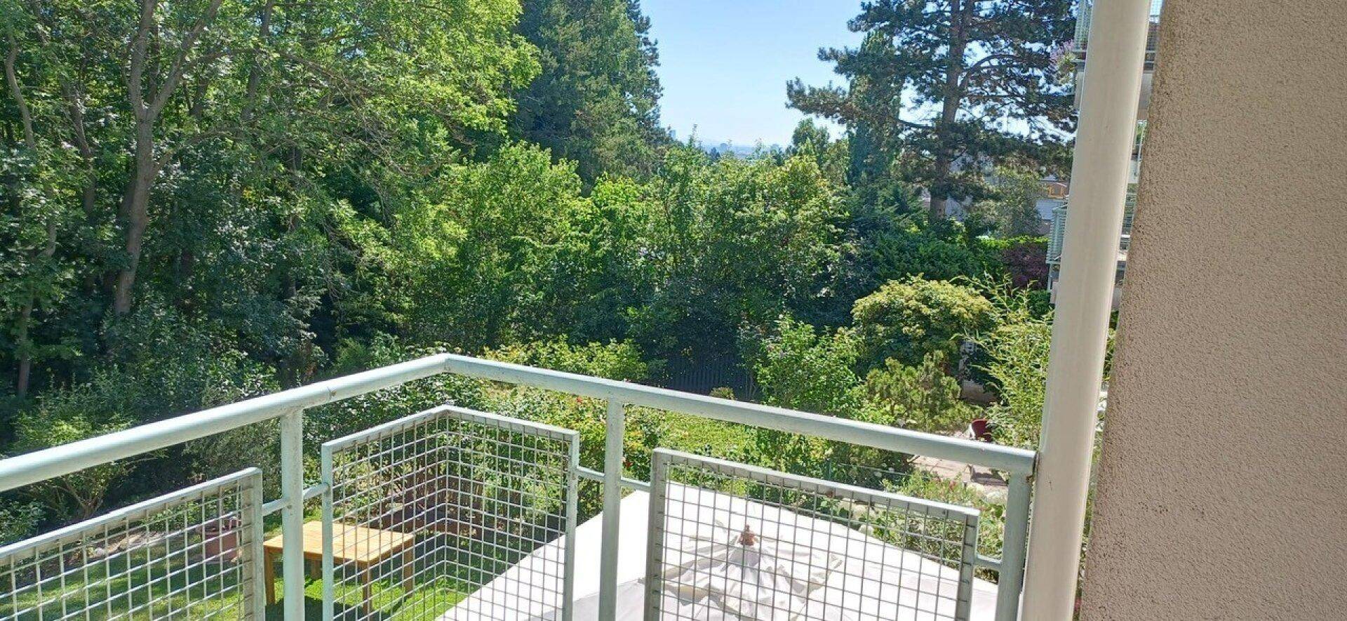 Weiterer Blick vom Balkon auf die grüne Umgebung und die Baumwipfel unter blauem Himmel.
