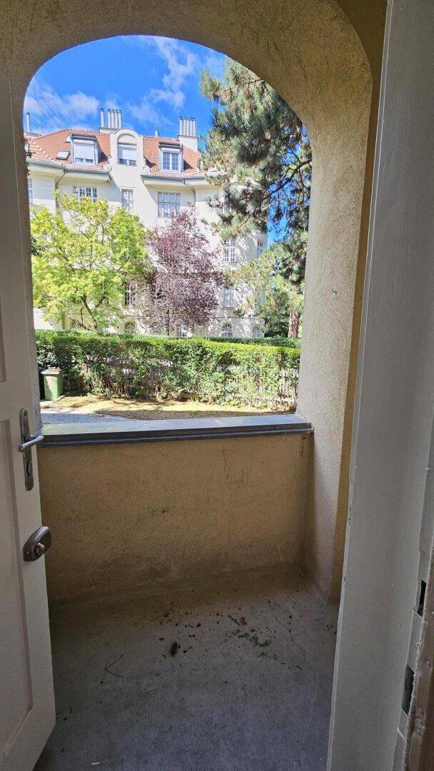 Überdachte Loggia mit Blick auf ein gegenüberliegendes Gebäude und grüne Hecken.