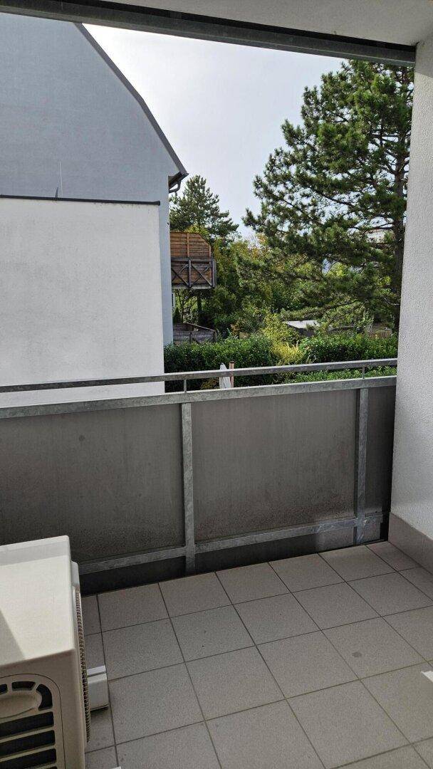 Kleiner Balkon mit Fliesenboden und Blick auf Bäume und Nachbargebäude.