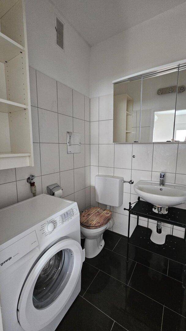 Badezimmer mit Waschmaschine, WC, Waschbecken und weißen Wandfliesen.