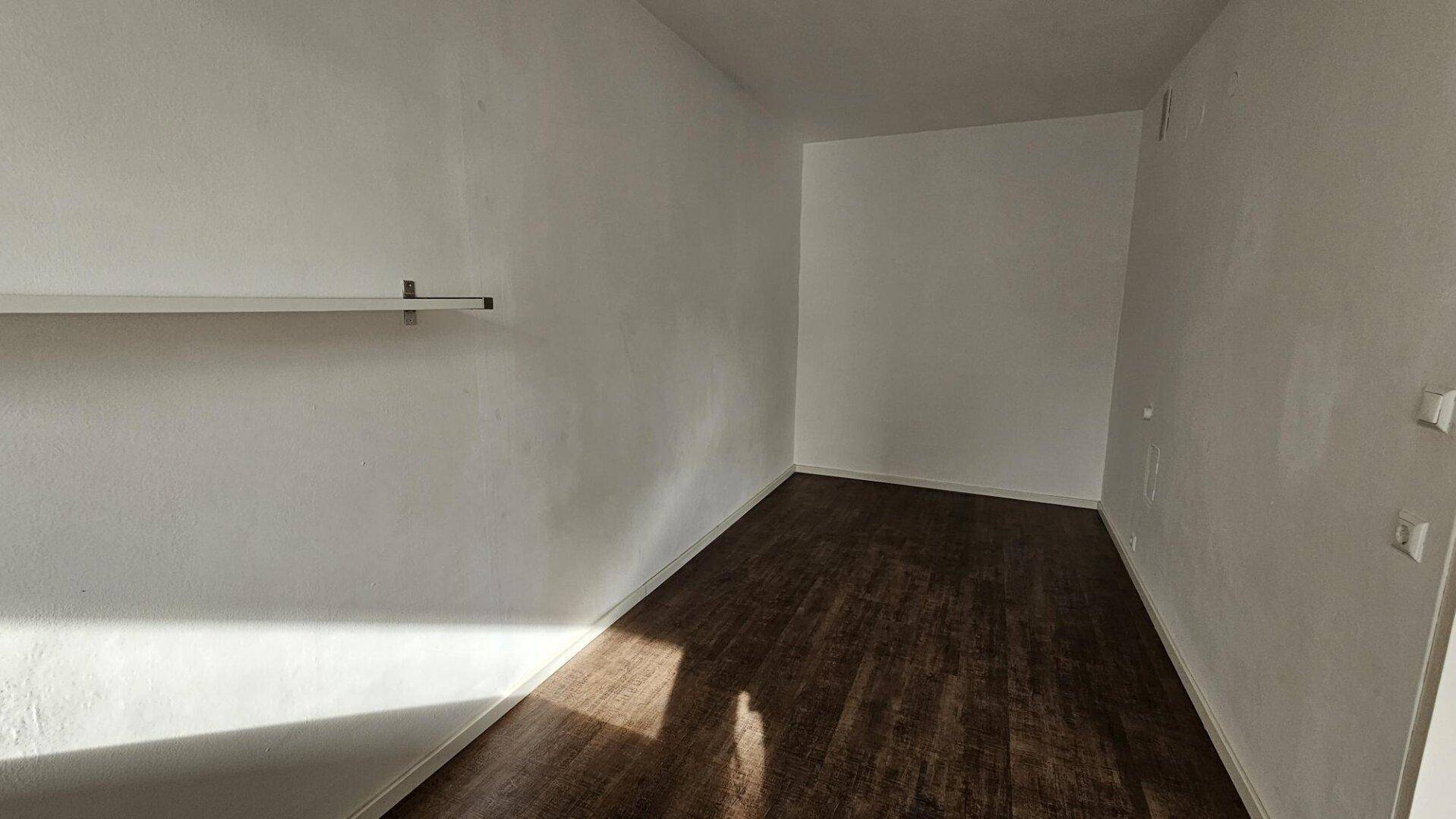 Leerer Raum mit weißen Wänden und dunklem Laminatboden, bereit zur individuellen Gestaltung.
