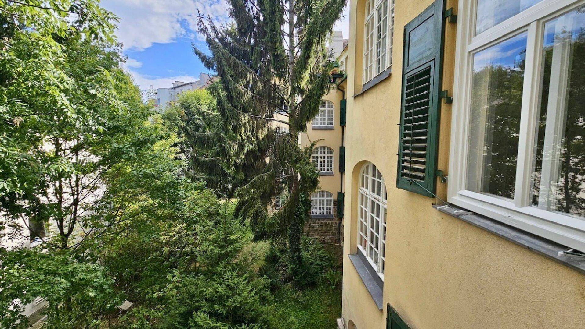 Blick aus dem Fenster auf eine charmante gelbe Fassade mit grünen Fensterläden und üppigem Baumbestand.
