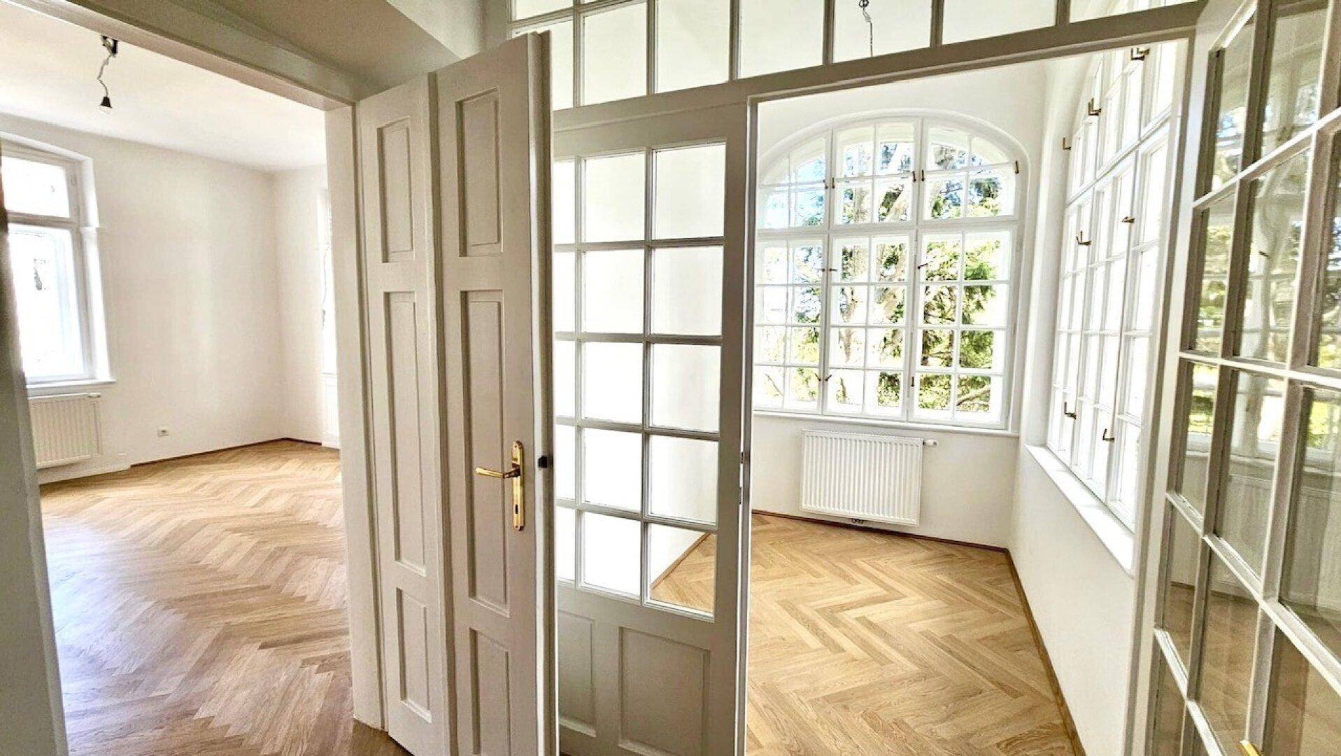 Blick durch eine Flügeltür in ein helles Zimmer mit großen Bogenfenstern und Fischgrätparkett.