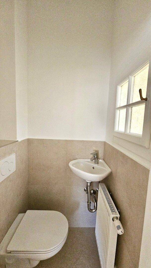 Kleines Gäste-WC mit Wand-WC, Eckwaschbecken, Heizkörper und einem kleinen Fenster.