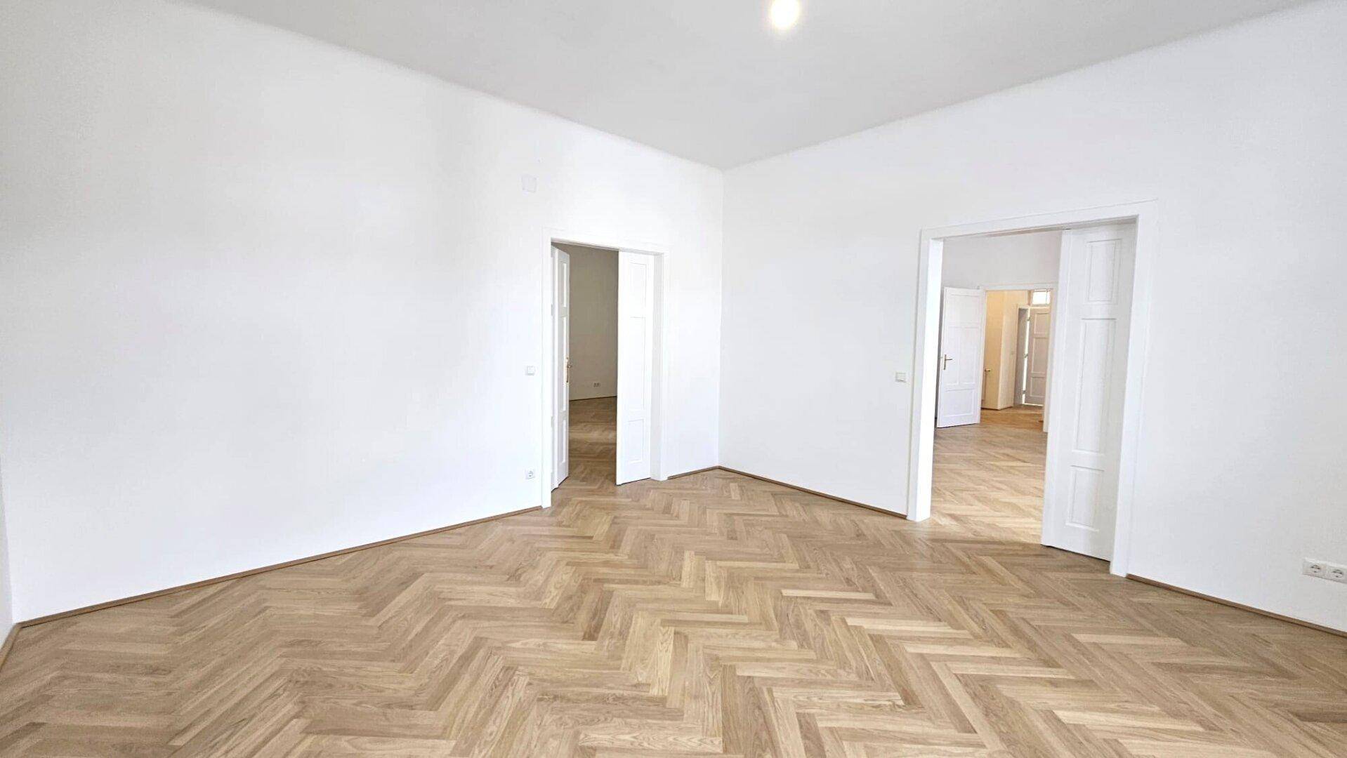 Geräumiges Zimmer mit Fischgrätparkett und mehreren Türen, die verschiedene Bereiche verbinden.