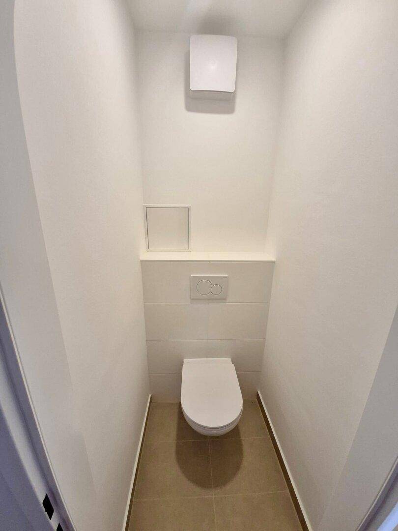 Kleines, separates WC mit wandhängender Toilette und einem Lüftungsventil an der Decke.