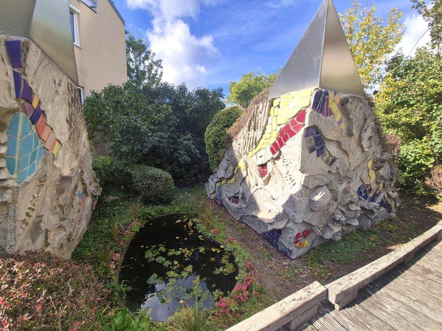 Kreativer Gartenbereich mit Mosaik-Skulpturen und einem kleinen Teich unter blauem Himmel.