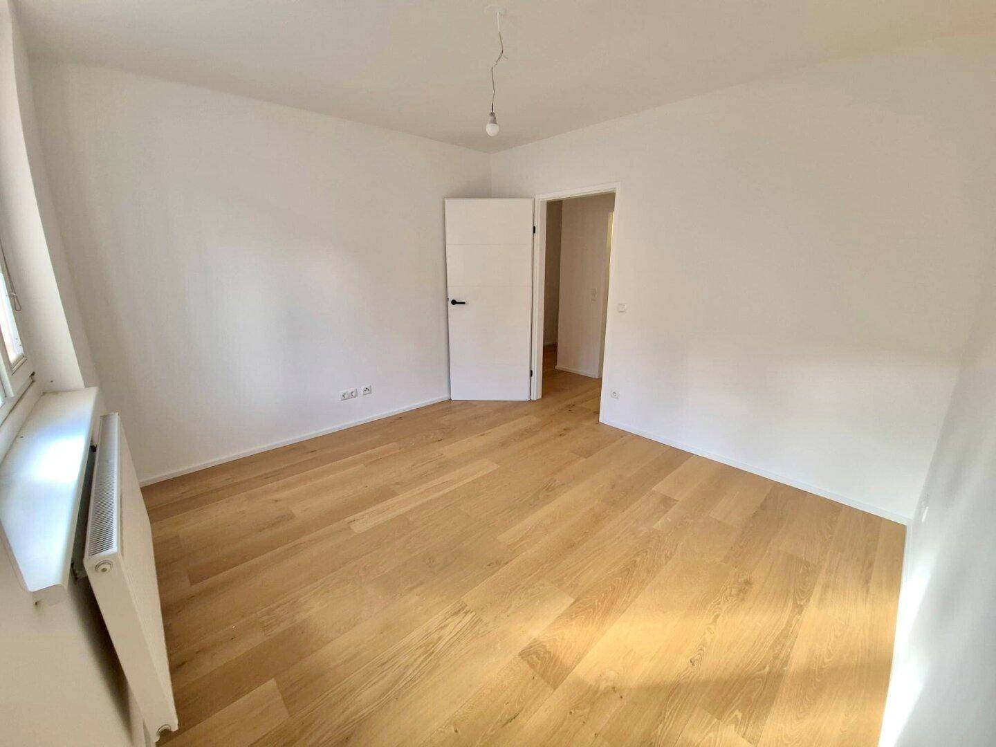 Heller Raum mit Holzboden und einer weißen Tür, die zu einem angrenzenden Bereich führt.