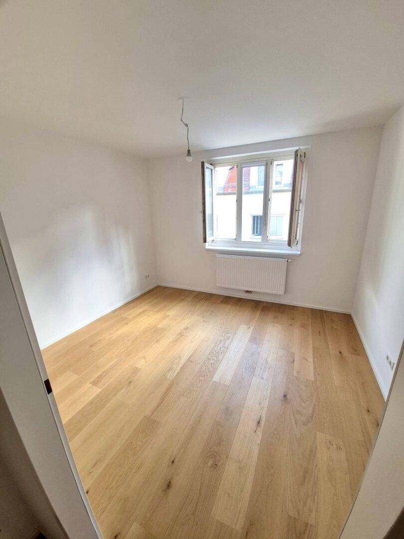 Helles, leeres Zimmer mit neuem Holzparkettboden und einem Fenster mit Heizkörper darunter.