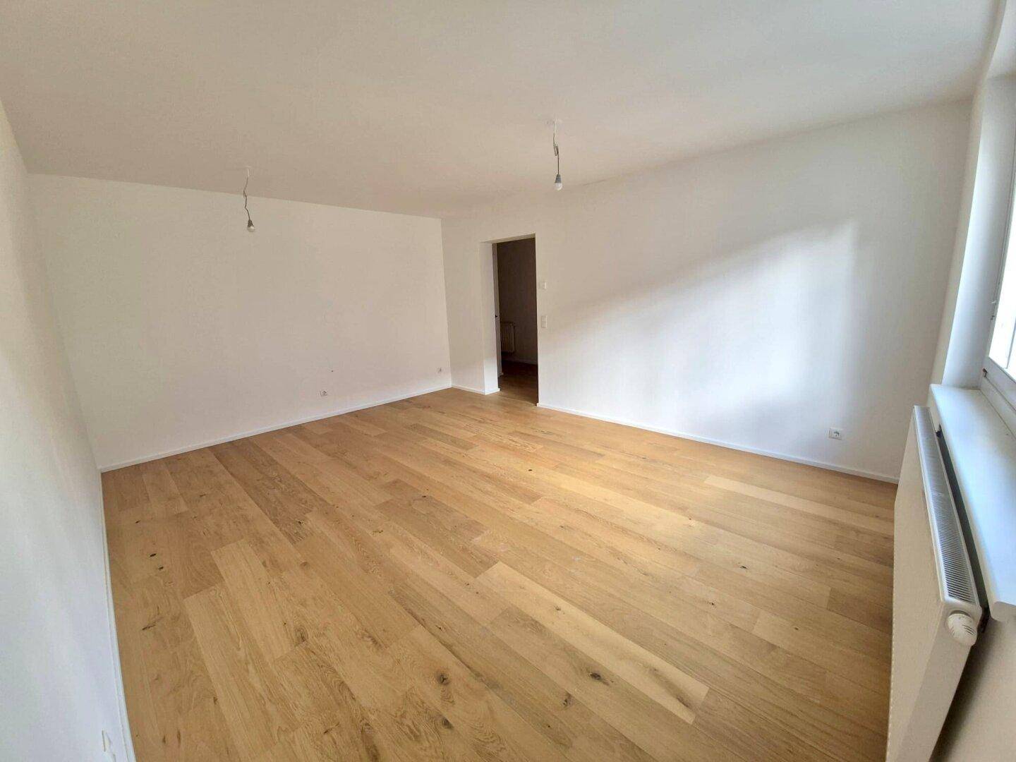 Leeres Zimmer mit hellem Holzparkettboden und einem offenen Durchgang zu einem weiteren Raum.