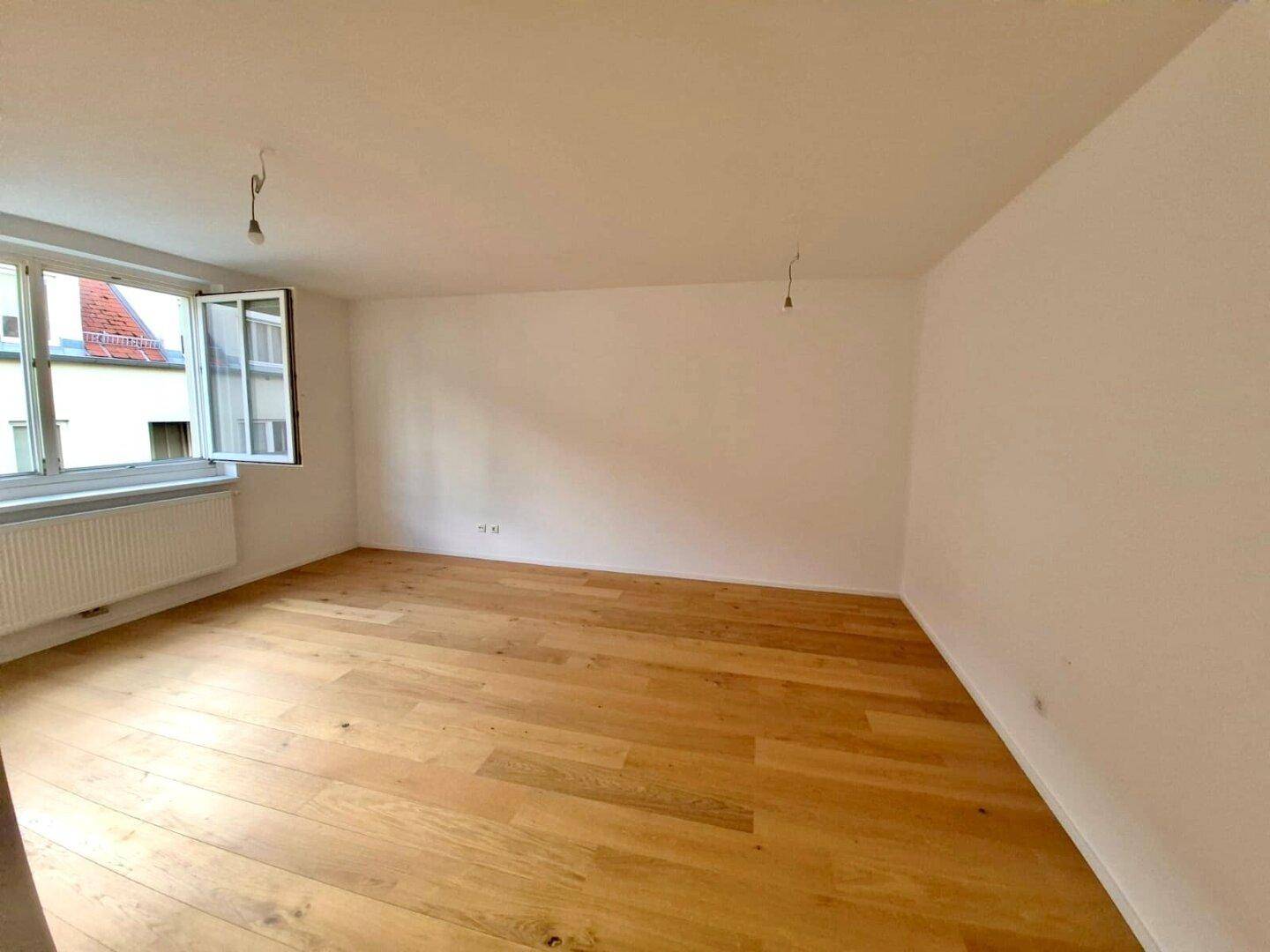 Geräumiges, helles Zimmer mit Holzboden und einem Fenster, das viel Tageslicht hereinlässt.