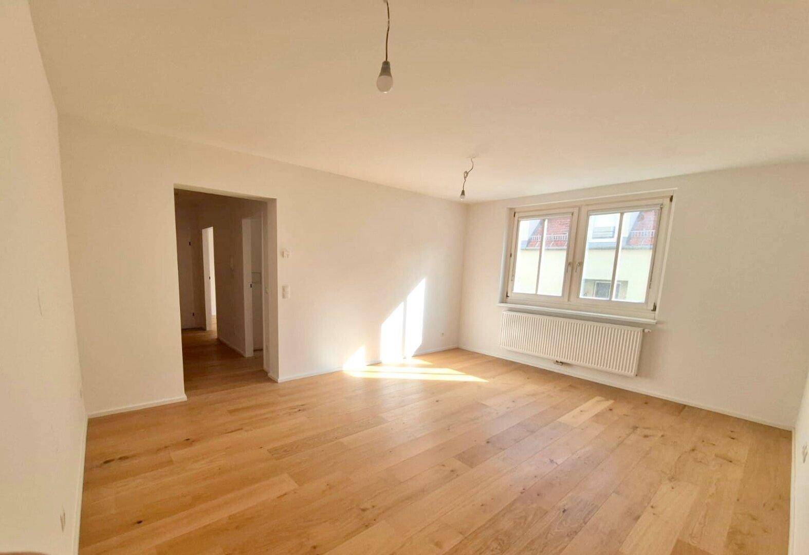 Großes, helles Zimmer mit Holzboden, einem Fenster und einem offenen Zugang zum Flur.