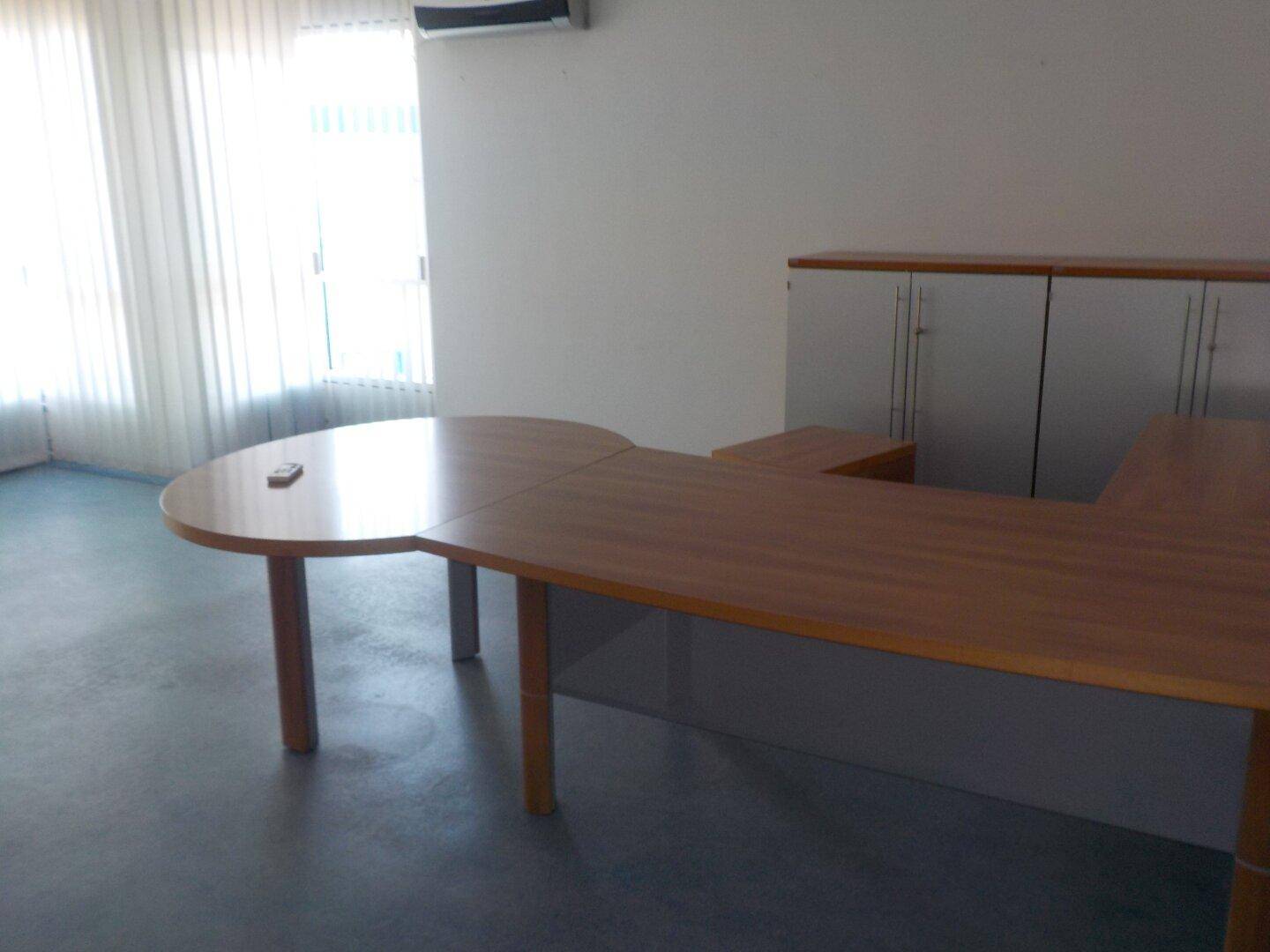 Büroraum mit einem geschwungenen Holztisch und einem weißen Schrank, gut beleuchtet.