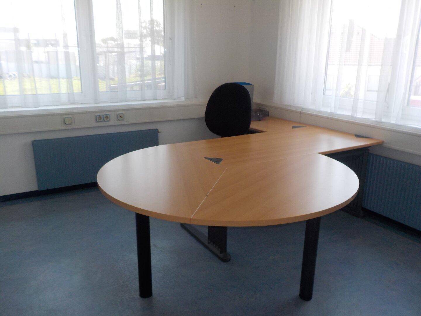 Büroraum mit einem runden Holztisch, der an einen Eckschreibtisch anschließt, und einem Bürostuhl.
