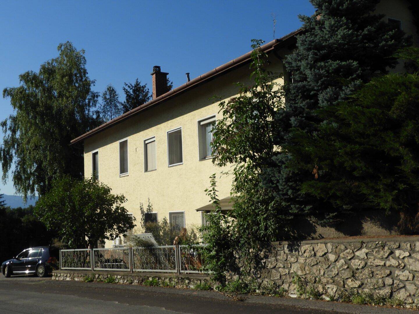 Seitenansicht des Wohnhauses mit gelber Fassade und umgebender Vegetation unter blauem Himmel.