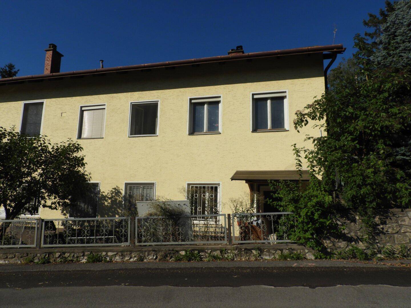 Vorderansicht des Wohnhauses mit gelber Fassade, Fenstern und einem kleinen Vordach im Sonnenlicht.