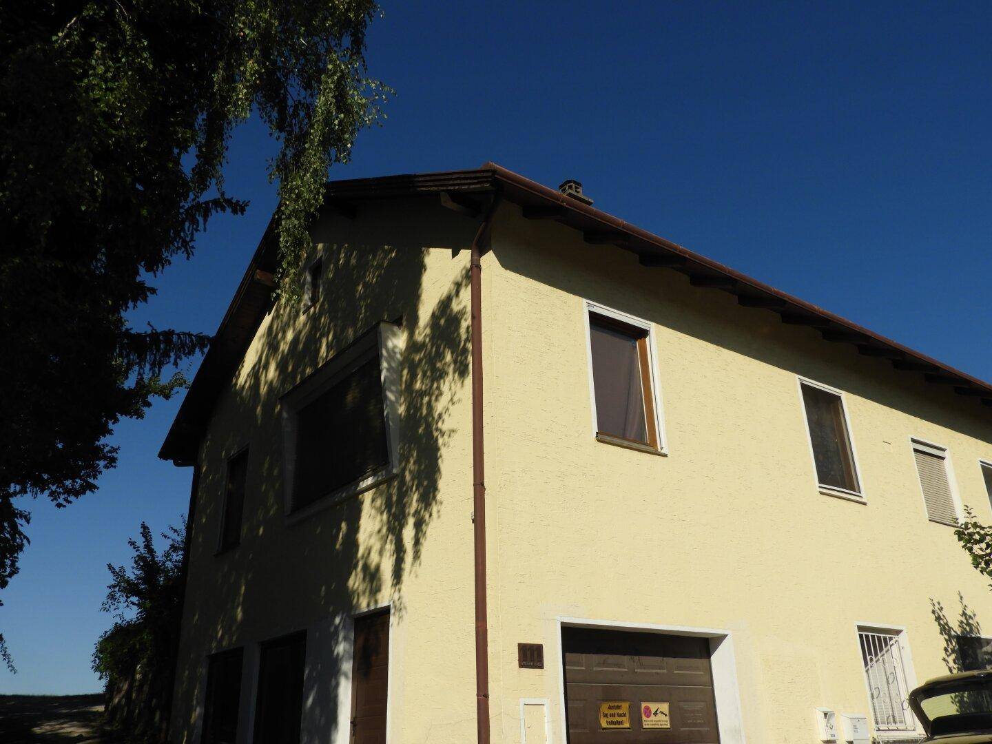 Eckansicht des Wohnhauses mit gelber Fassade, Garagentor und Fenstern unter klarem blauen Himmel.