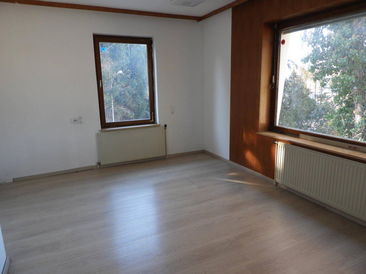 Helles Zimmer mit Laminatboden, zwei Fenstern und Heizkörpern, bereit zur individuellen Einrichtung.