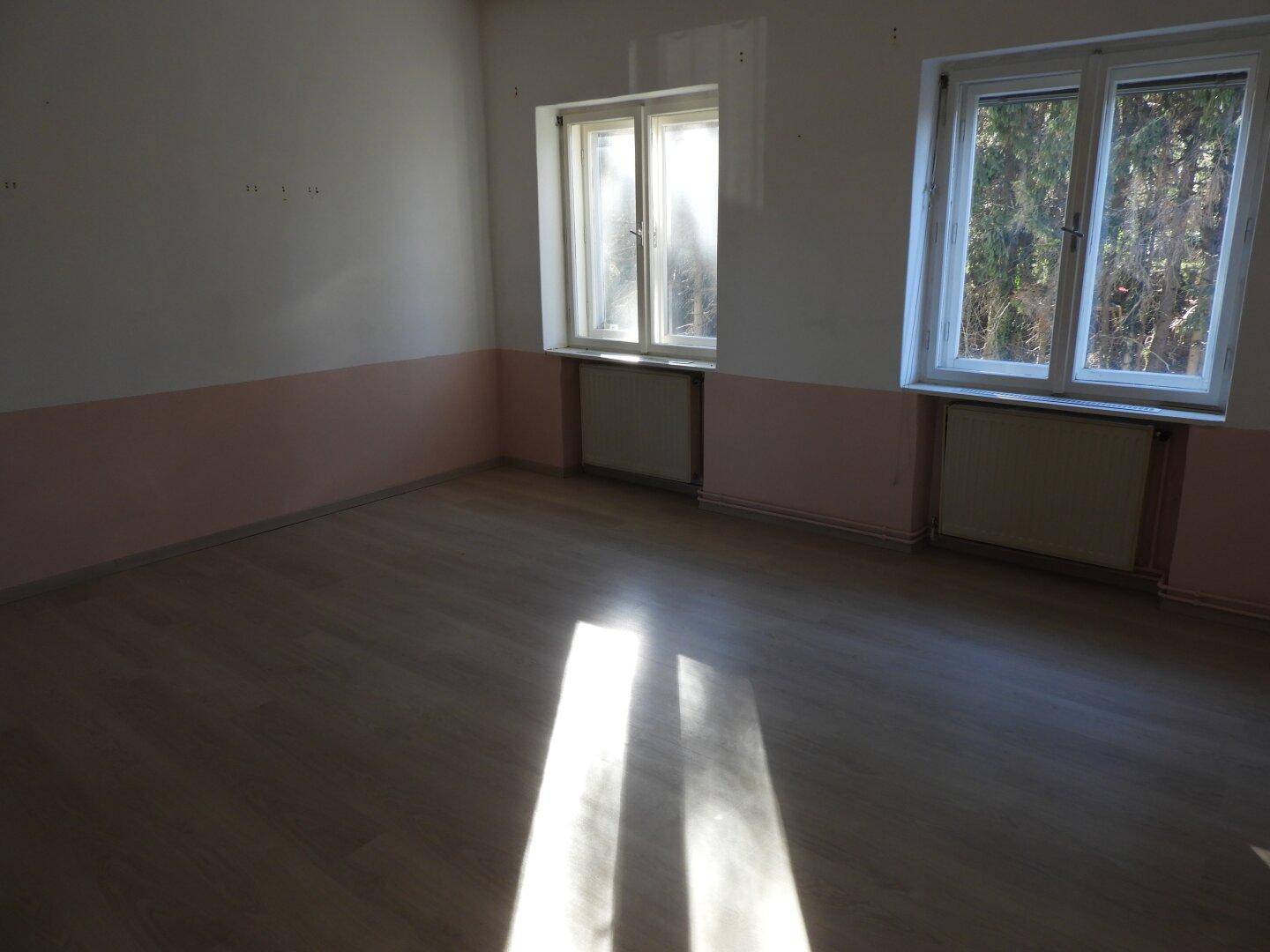 Helles Zimmer mit Laminatboden und zwei Fenstern, durch die Sonnenlicht auf den Boden fällt.