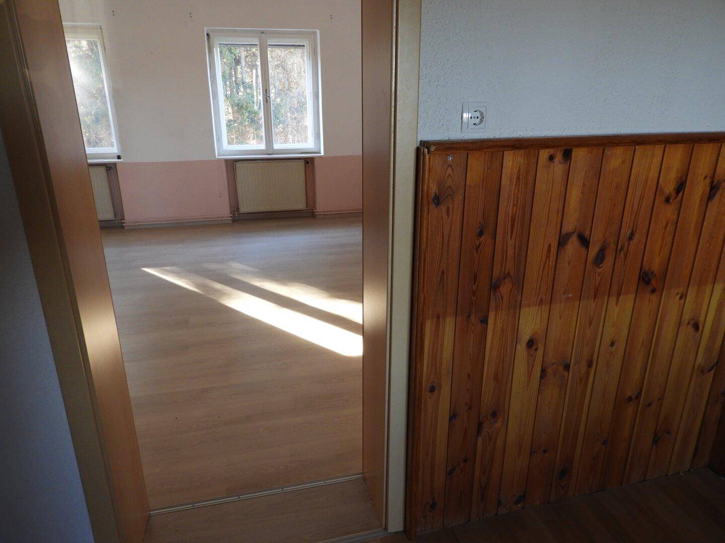 Türrahmen führt in ein helles Zimmer mit Laminatboden und Fenstern, die Sonnenlicht hereinlassen.