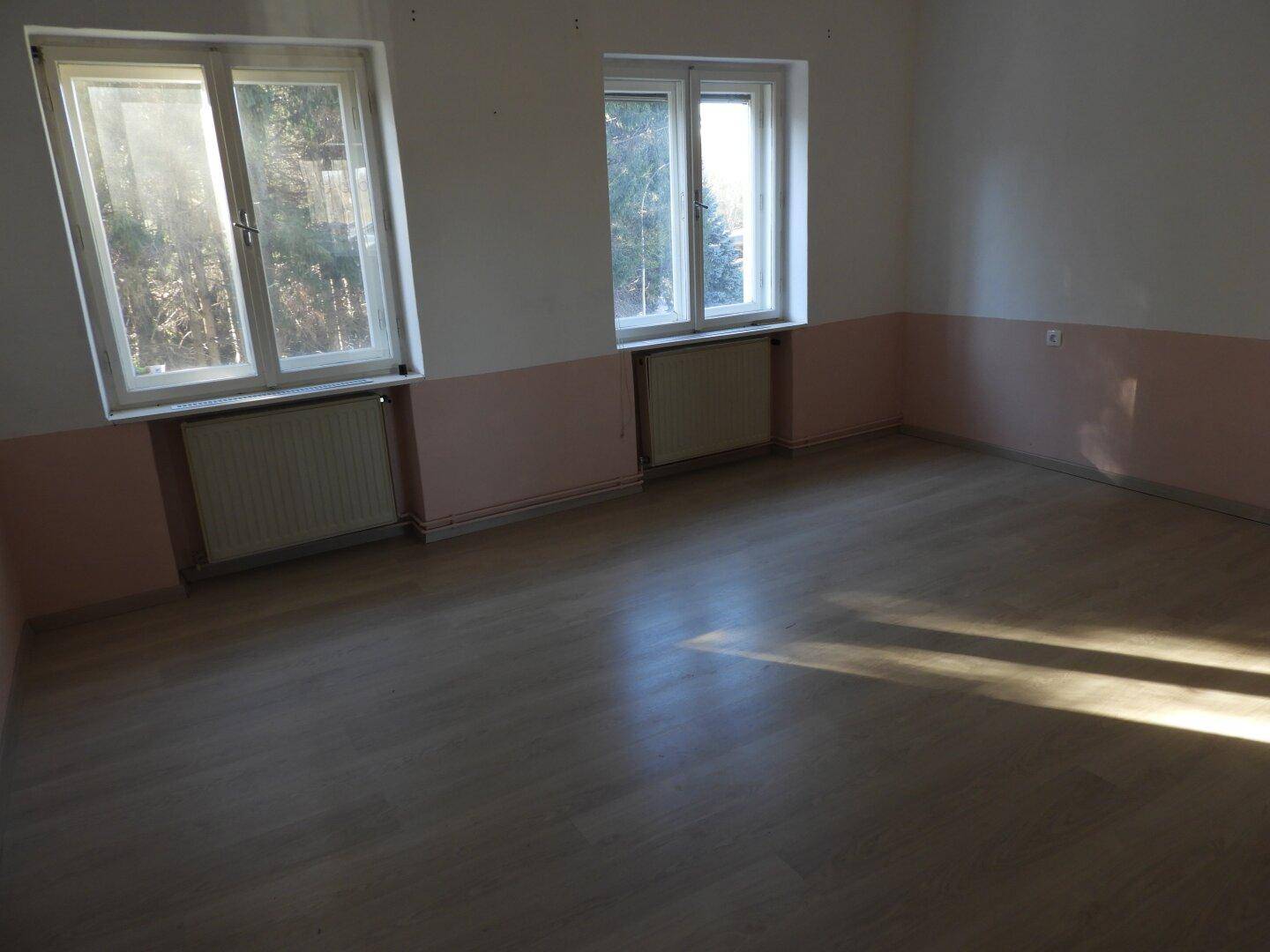 Helles Zimmer mit Laminatboden und zwei Fenstern, durch die viel Sonnenlicht einfällt.