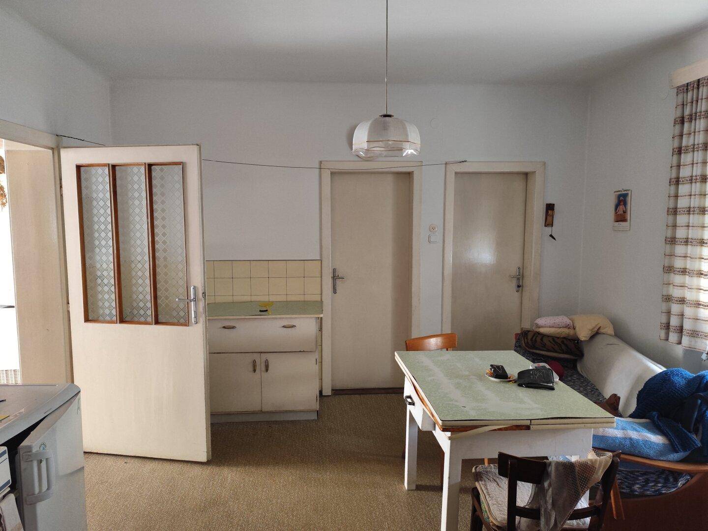 Einfache Küche mit altem Tisch, kleiner Anrichte und zwei Türen in einem älteren Gebäude.