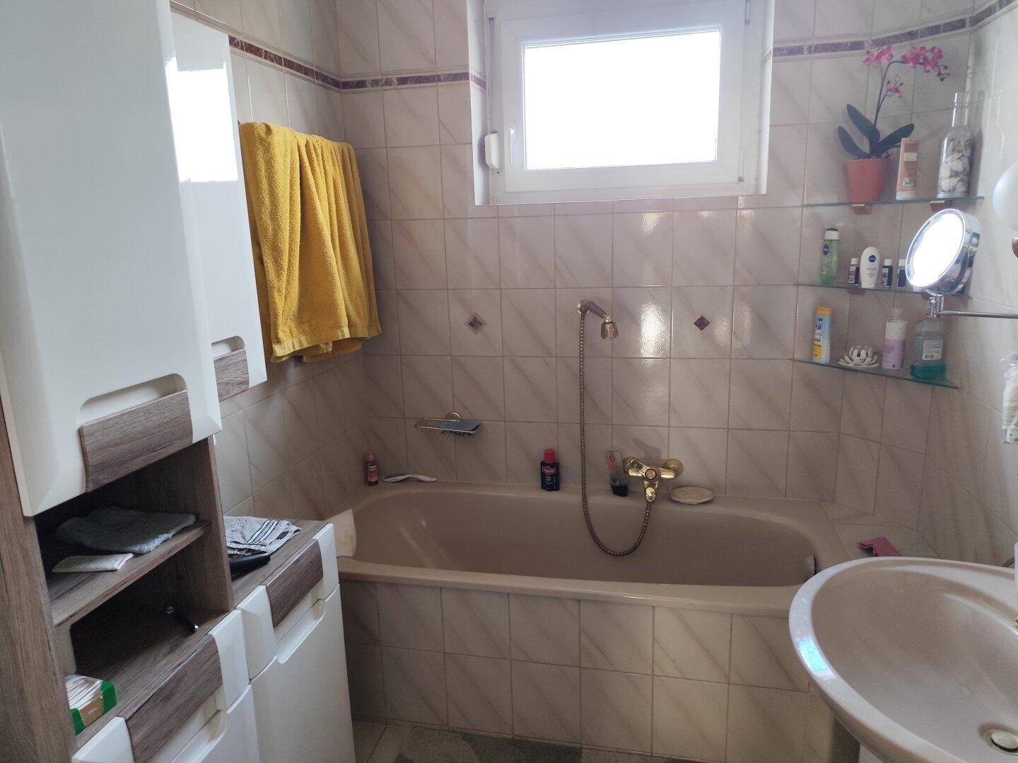 Badezimmer mit Badewanne, Waschbecken und Fenster, ausgestattet mit hellen Fliesen.