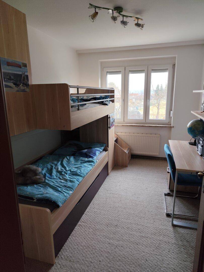 Kinderzimmer mit Etagenbett, Schreibtisch und Teppichboden, hell durch großes Fenster.