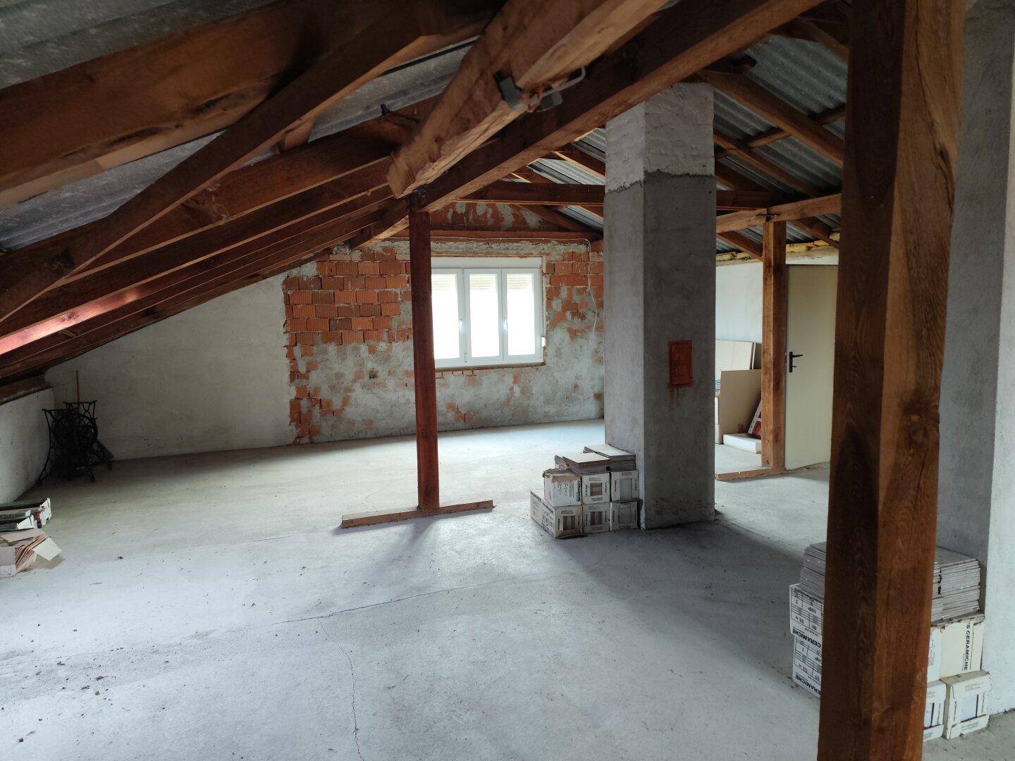 Unfertiger Dachboden mit sichtbaren Holzbalken, Betonboden und Fenster, bereit für den Ausbau.