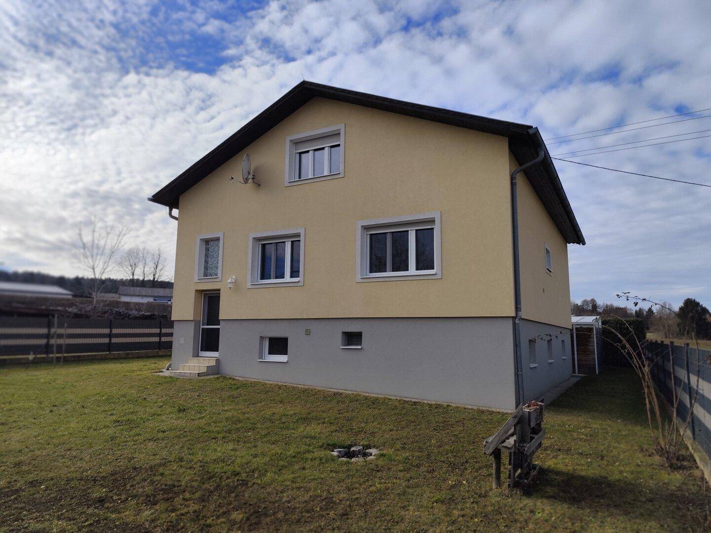 Seitenansicht des Hauses mit gelber Fassade und grauer Sockelzone unter bewölktem Himmel.