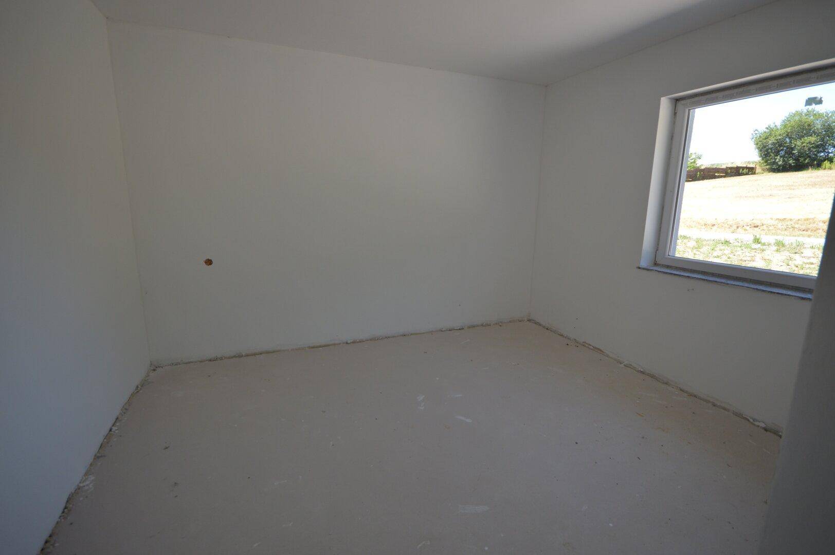 Helles Schlafzimmer im Rohbau mit einem Fenster, das viel Tageslicht und eine grüne Aussicht bietet.