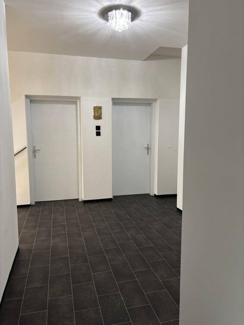 Heller Flur mit dunklen Fliesen und weißen Türen, beleuchtet von einer Deckenleuchte.