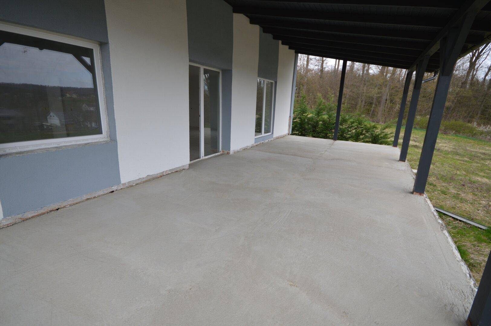 Unfertige Beton-Terrasse mit Zugang zum Innenbereich und Blick in die Natur.