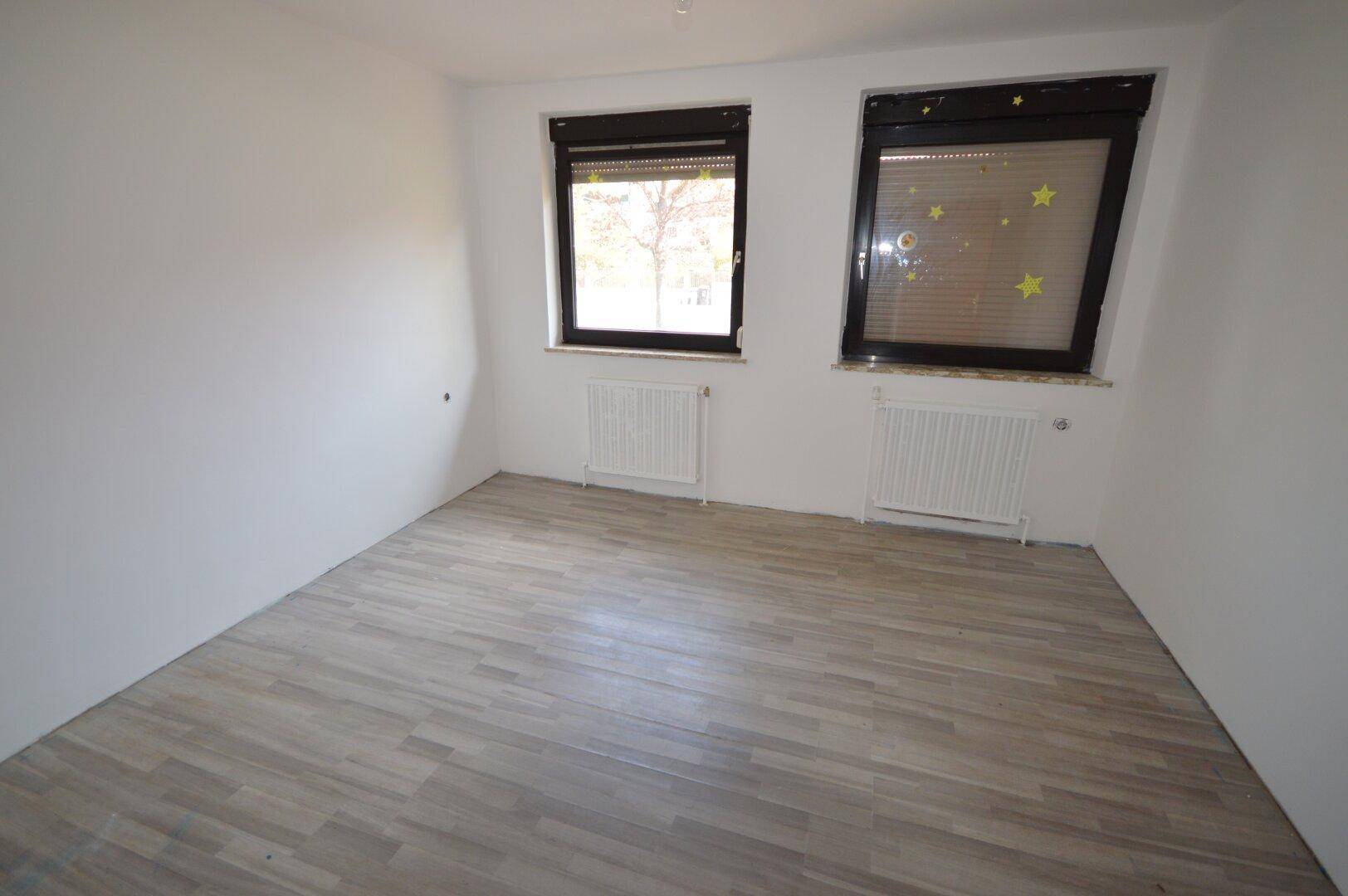 Schlafzimmer mit hellem Laminatboden und zwei Fenstern, die viel Tageslicht hereinlassen.