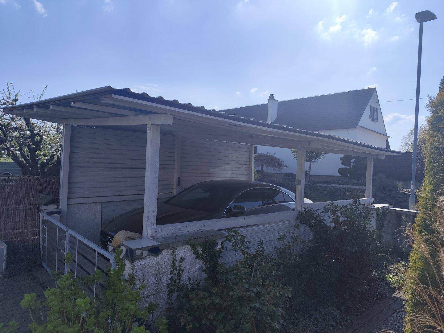 Überdachter Carport mit einem geparkten Auto und einem weiteren Haus im Hintergrund.