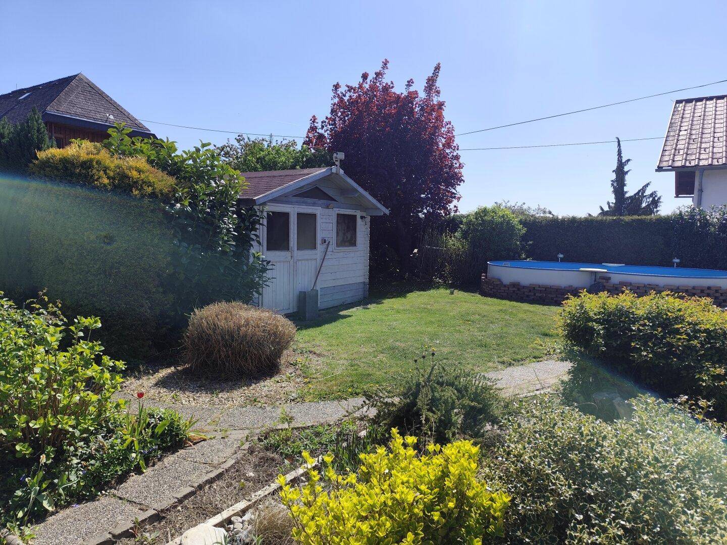 Gepflegter Garten mit Gartenhaus, Rasenfläche und vielfältiger Bepflanzung unter blauem Himmel.