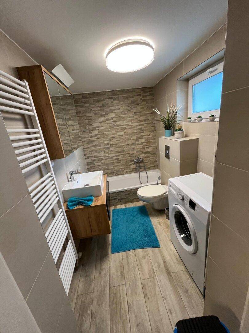 Modernes Badezimmer mit Badewanne, Waschbecken, WC und Waschmaschine auf Holzoptik-Fliesen.