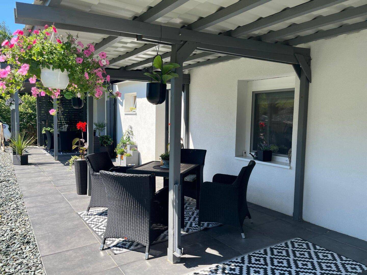 Überdachte Terrasse mit schwarzen Gartenmöbeln und Blumenampeln, ideal für entspannte Stunden.