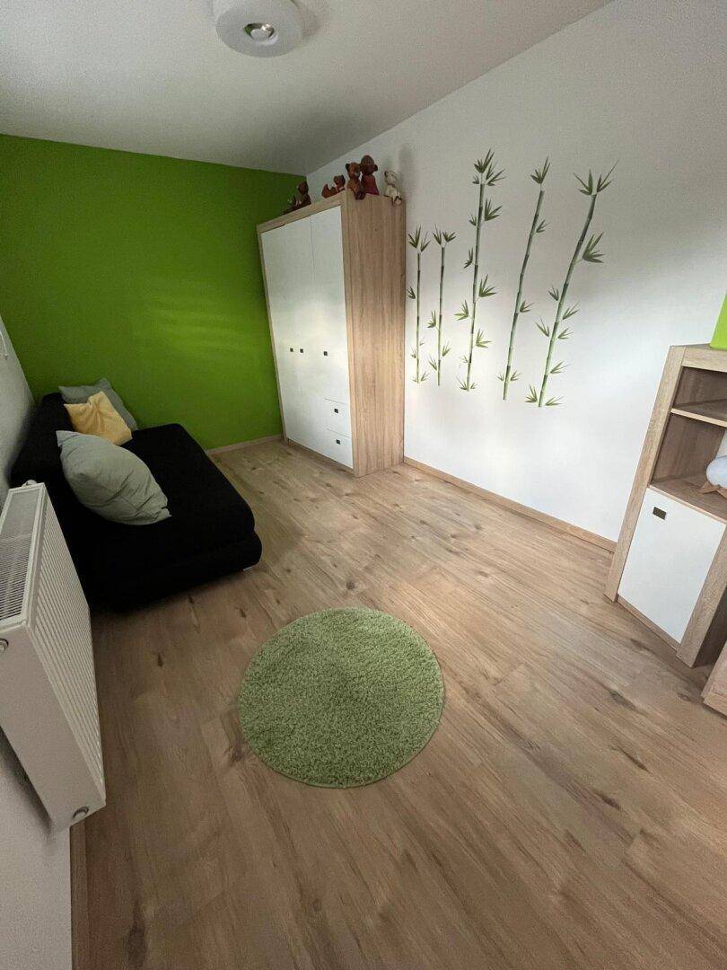Kinderzimmer mit grüner Akzentwand, Holzmöbeln und Laminatboden, hell und freundlich gestaltet.