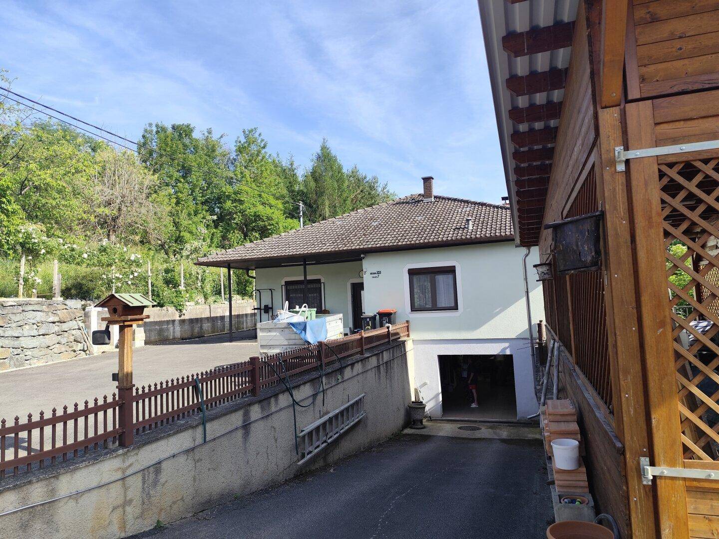 Hausfassade mit Garagenzufahrt und angrenzender Holzkonstruktion, umgeben von Natur.