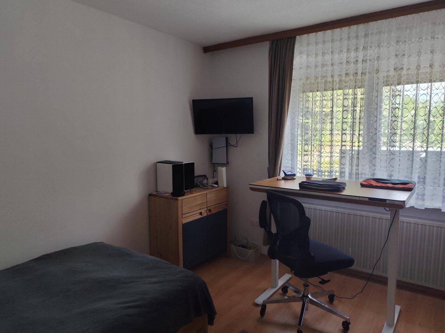 Helles Schlafzimmer mit Schreibtisch, Fernseher und Fenster mit Blick ins Grüne.