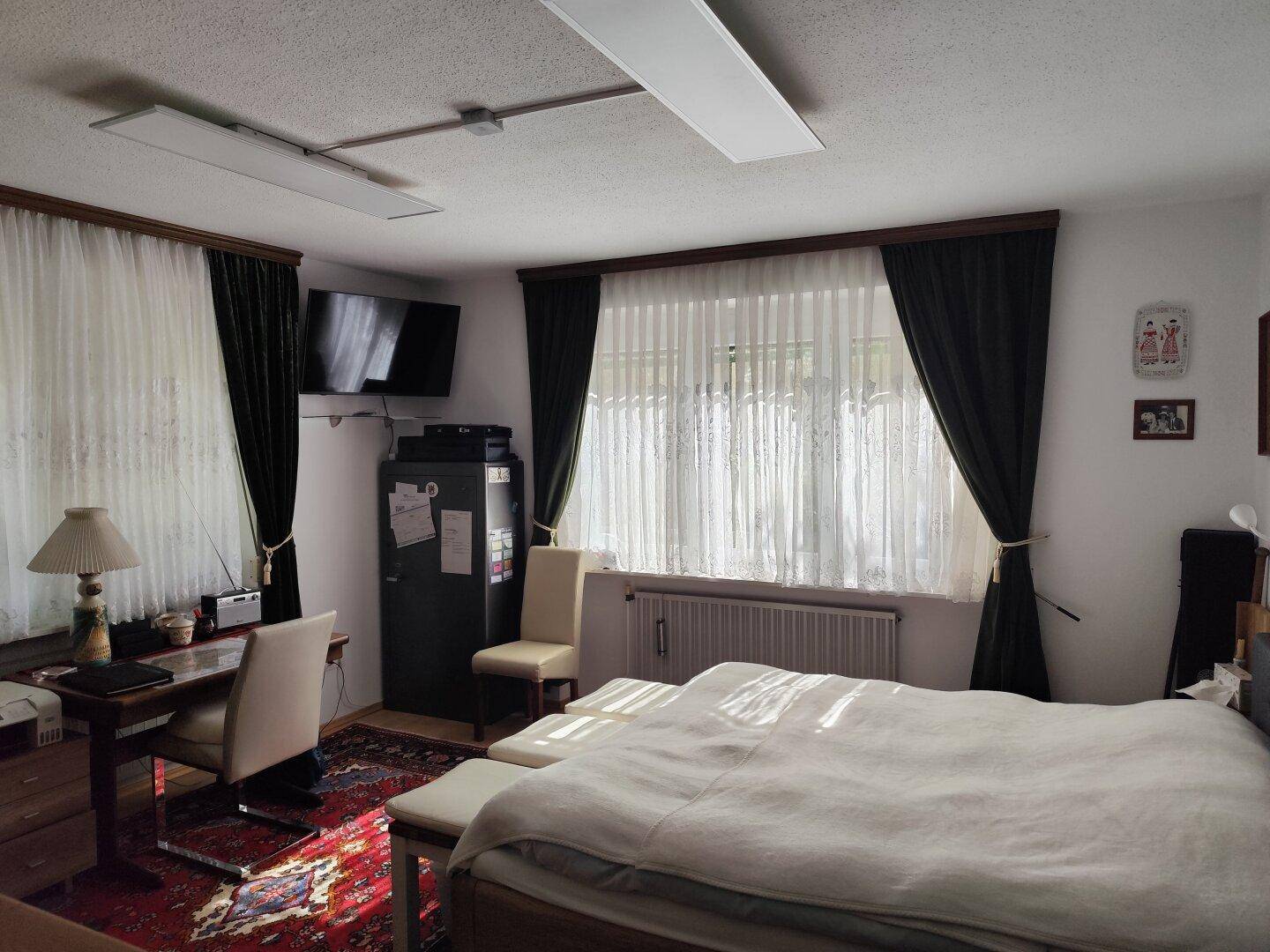 Schlafzimmer mit großem Bett, Schreibtisch und Blick ins Grüne durch die Fenster.