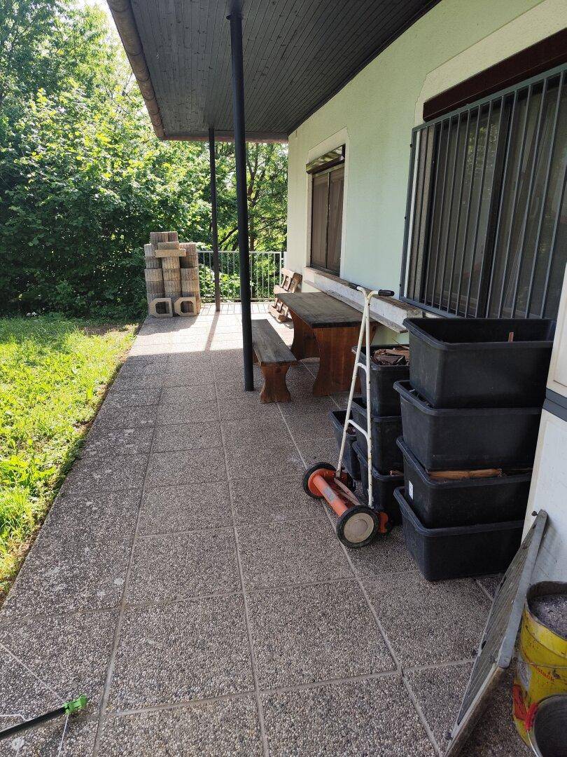 Überdachte Terrasse mit Fliesenboden, Holztisch und Bank, umgeben von viel Grün.