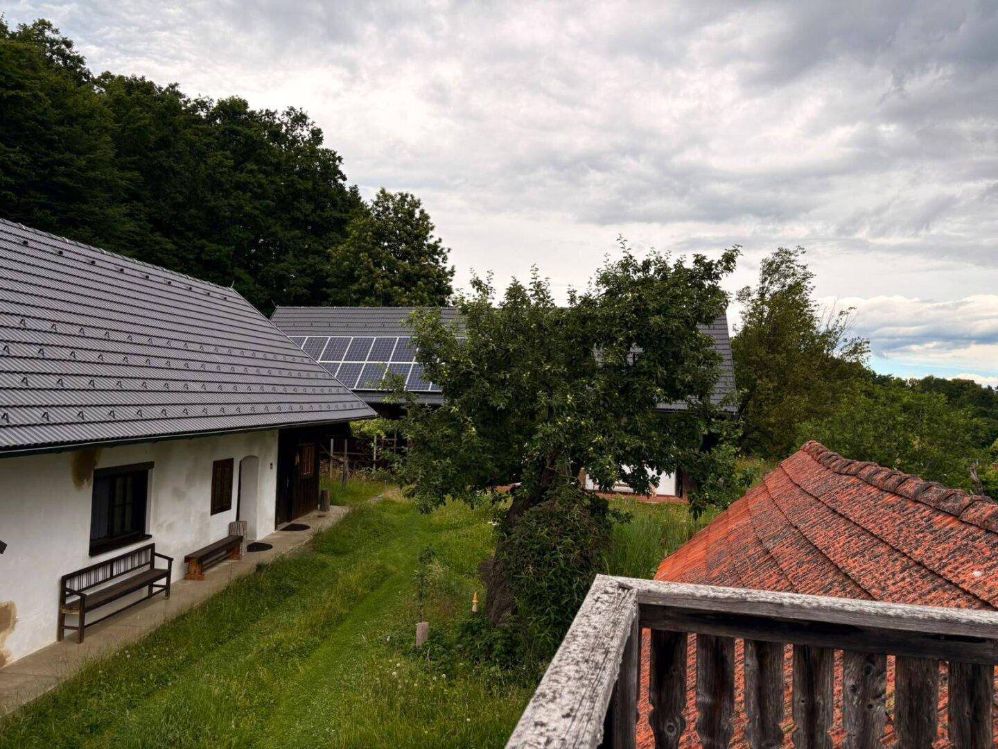Blick von einer Holzterrasse auf ein weißes Haus mit Photovoltaikanlage und grüner Umgebung.