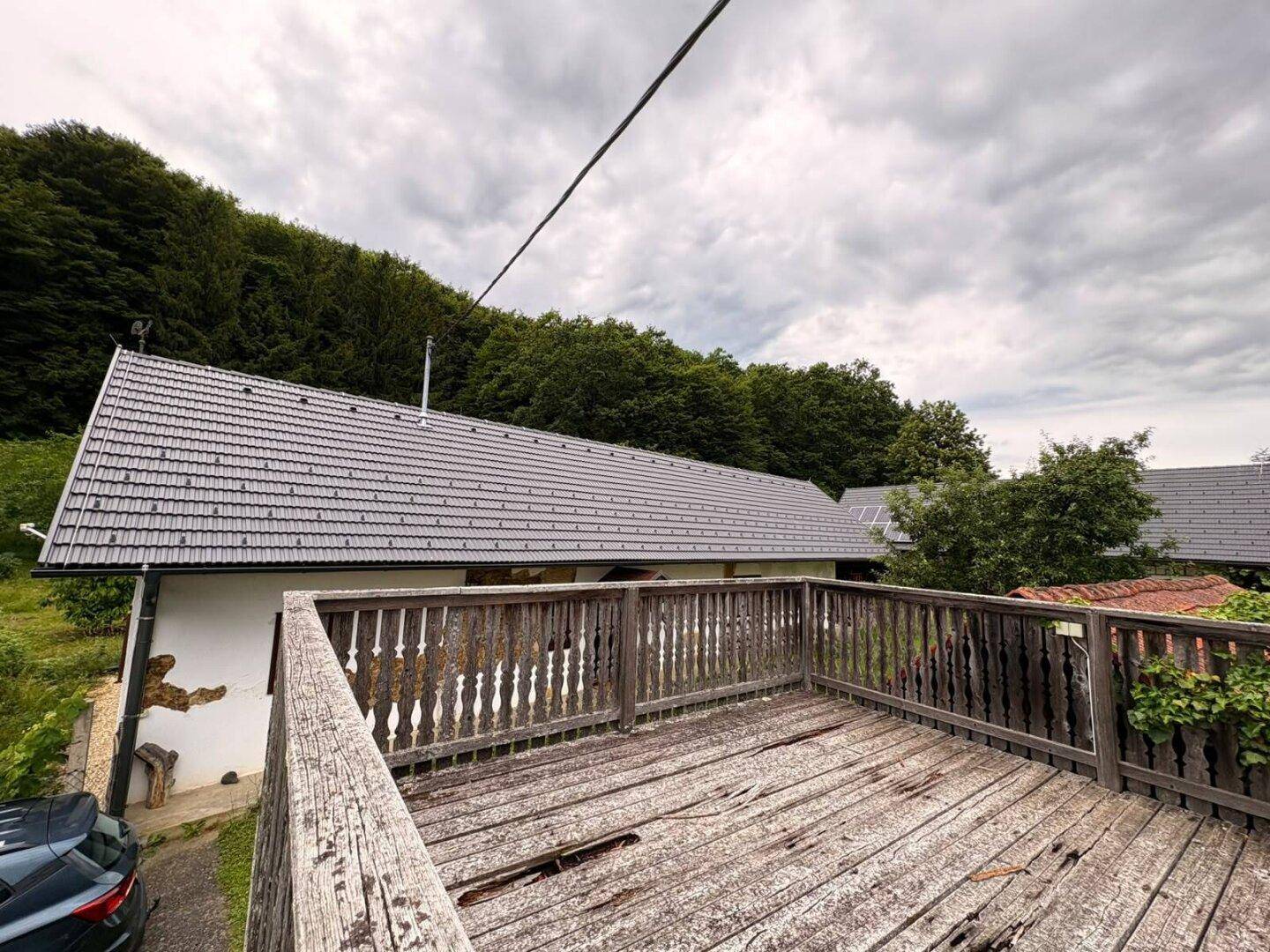 Blick von einer rustikalen Holzterrasse auf die Seite eines weißen Hauses mit grauem Ziegeldach.