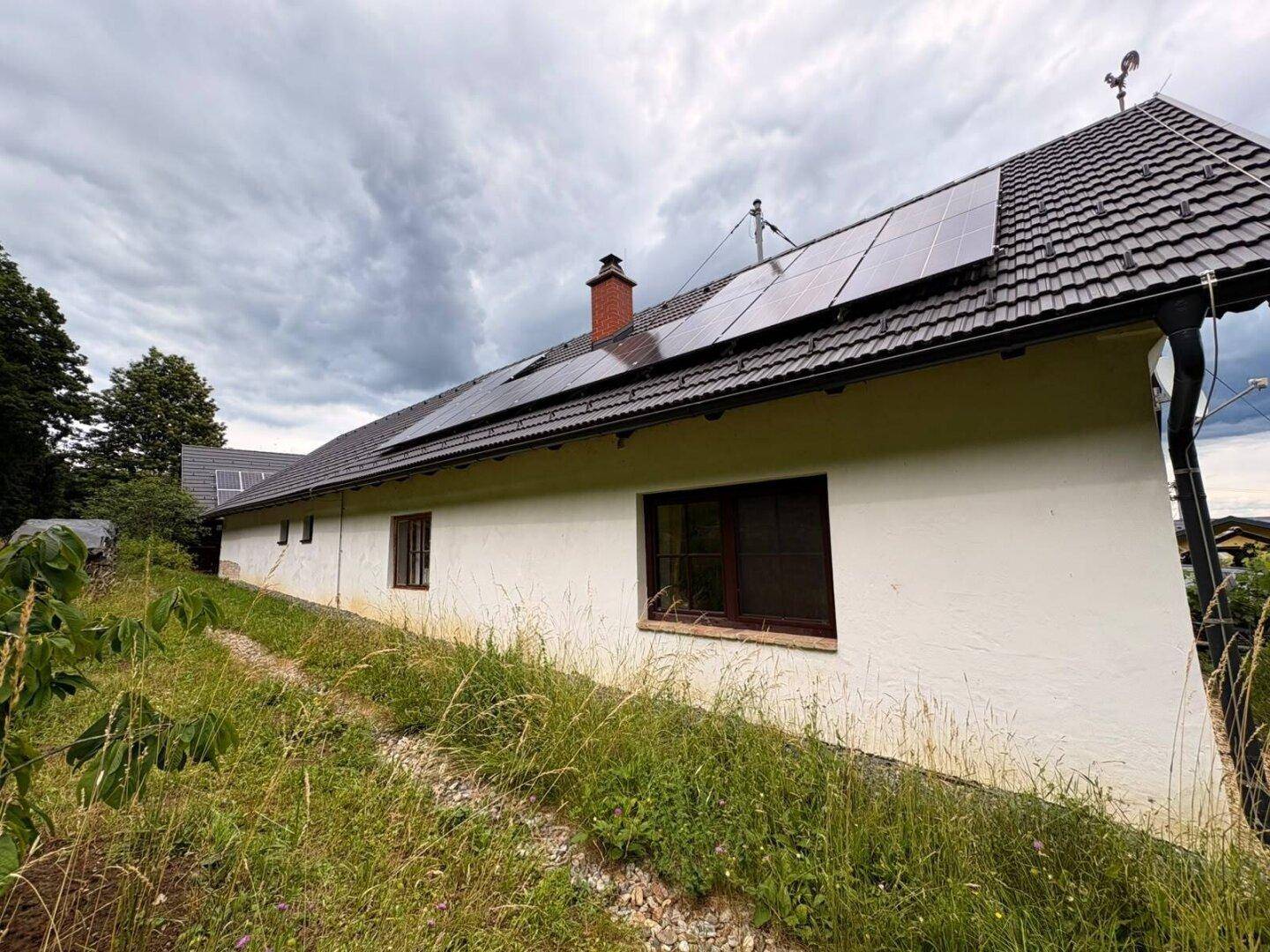 Seitenansicht eines weißen Hauses mit grauem Dach und Solaranlage auf einem grasbewachsenen Hang.